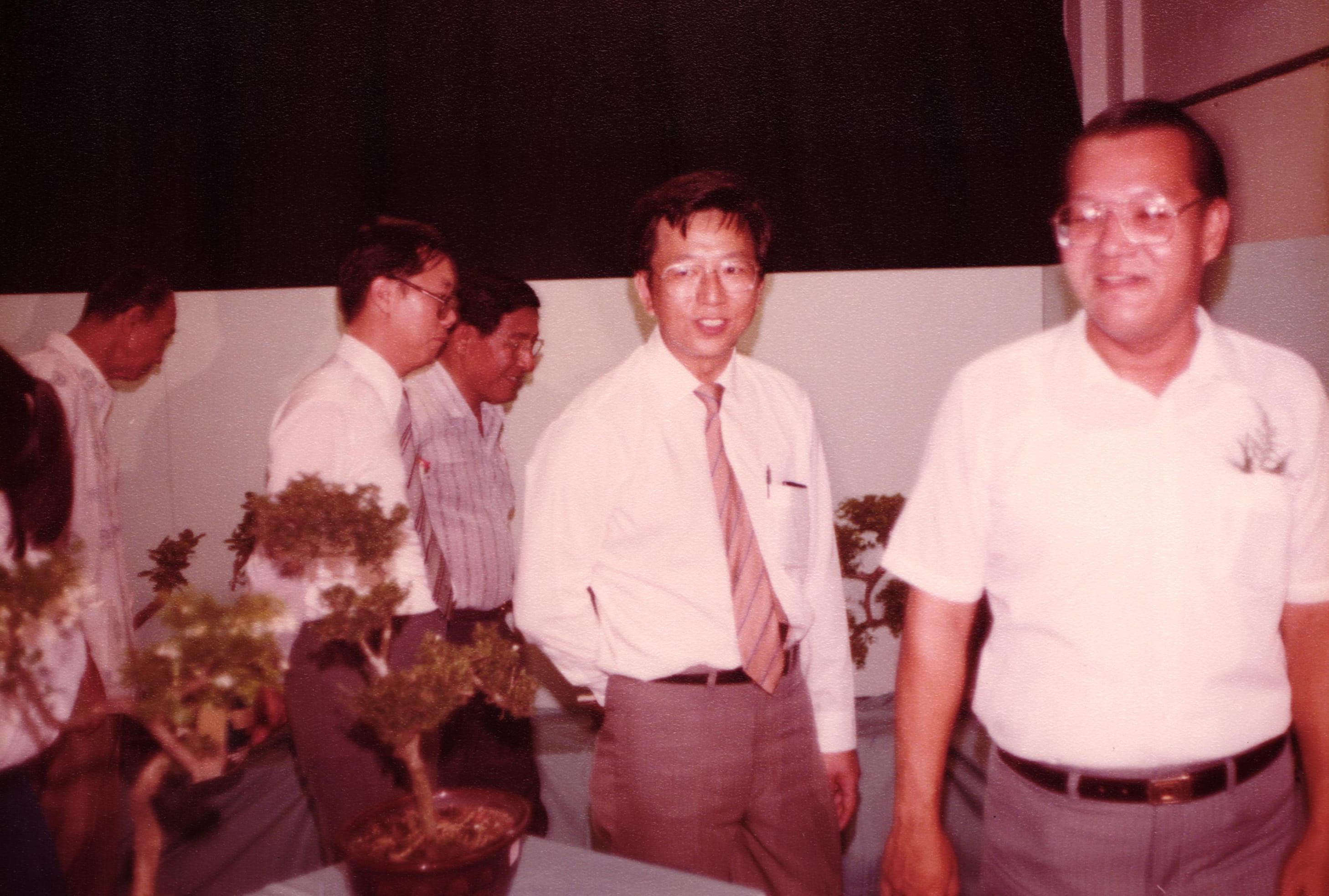 Several men in dress shirts stand near a display of small bonsai trees in a vintage indoor photo.